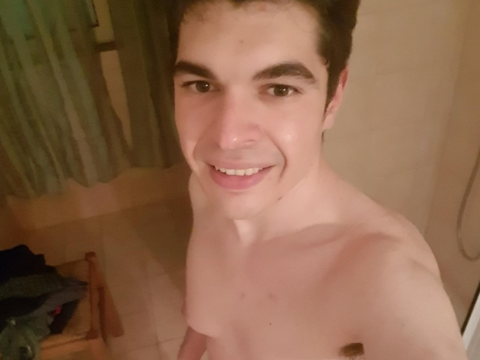 AdrianoX with a cute smile, short brown hair, and a slightly chubby, shirtless upper body, holding his small, flaccid, uncircumcised penis in a bathroom.