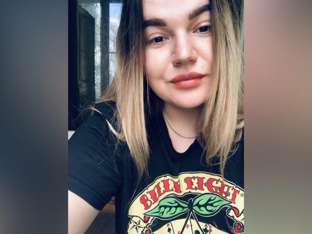 CherryXGirll, avec des cheveux gris-violets ondulés, un anneau au nez et un maquillage discret, portant un haut blanc, regardant directement l'appareil photo avec un léger sourire confiant..