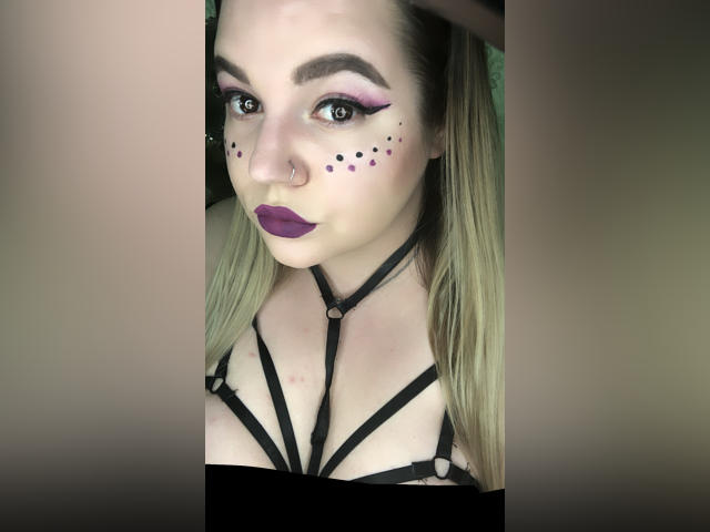 CherryXGirll, com seios grandes, vestindo lingerie preta, tem cabelos loiros longos e cacheados, tatuagens nos braços e está ajoelhada em uma cama com um cobertor branco fofo..