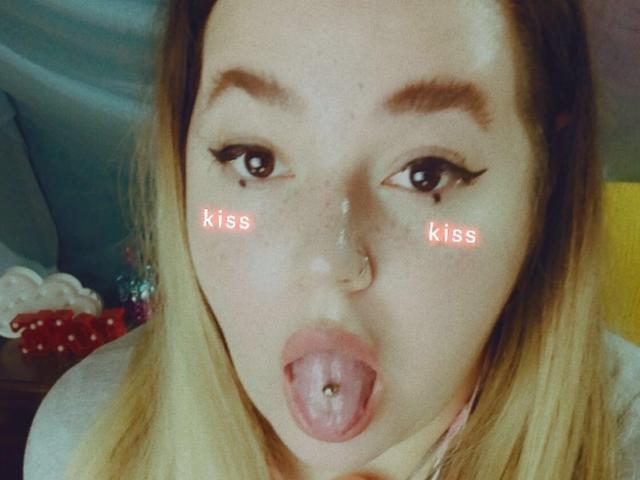 Os seios grandes e arredondados da CherryXGirll em um sutiã preto com alças finas, mostrando bastante o colo, com seu cabelo loiro cobrindo os ombros. Ela tem tatuagens nos braços..