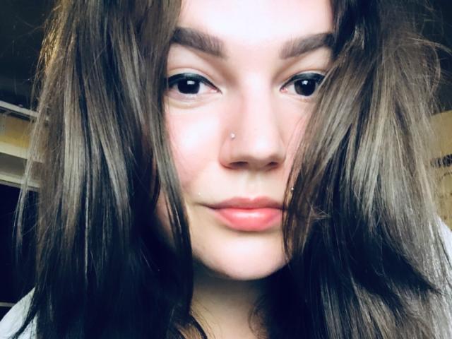 CherryXGirll's große, runde Brüste sind unter einem engen weißen Tanktop sichtbar, ihr blondes Haar fällt ihr herab, und auf dem Bild trägt sie schwarze Hosen..