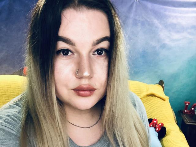 CherryXGirll heeft een schattig gezicht met bruine ogen, een neusring, een lip-piercing en een lichte glimlach. Haar bruine haar is los en ze draagt een rode armband..