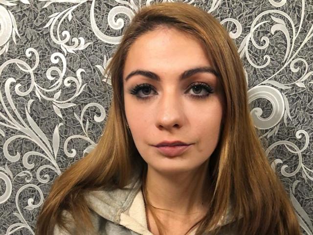 HotQueenAmelia with light brown hair, thick eyebrows, and subtle makeup, smiling slightly. She's wearing a white shirt, and her eyes are dark and slightly upturned.