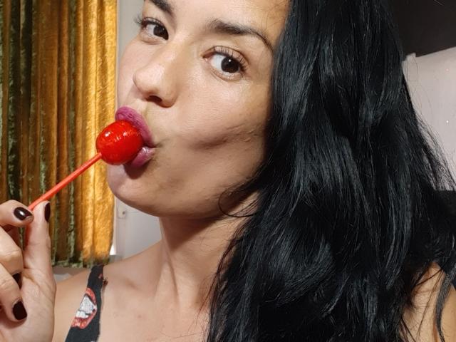 JuliaIce met zwart, golvend haar, blauwe ogen en roze lippenstift, draagt een paars kanten beha, zwarte gestreepte kousen, heeft haar arm omhoog en zit tegen een houten muur, waarbij haar borsten zichtbaar zijn..