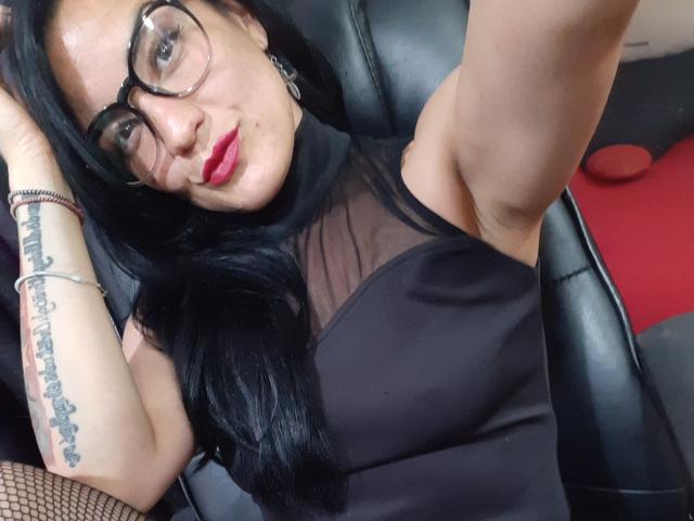 ValeryGirlLatin mit langen, schwarzen Haaren, heller Haut und rosafarbenem Lippenstift, die an einem roten Zuckeräpfel lutscht. Sie trägt ein schwarzes Tanktop mit einem roten Aufkleber..