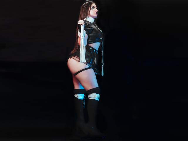 AbbyShiine with long brown hair, wearing black leather outfit, exposing her ass, black thigh straps, and knee-high boots, holding her shirt, standing in dark, blue-lit background.