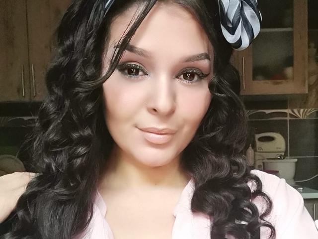 LexyRene has curly black hair, green eyeshadow, and a pink shirt. She's smiling with her hand on her hair, wearing a black and white striped headband.