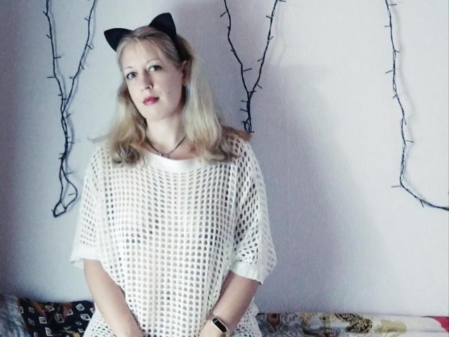 SoftGella met blond haar, die zwarte kattenoren draagt en een witte kanten top, zittend, met kleine borsten, lichte huid, rode lippenstift en een zwarte polsband, kijkt iets opzij..