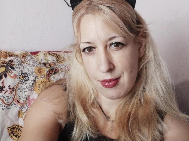 SoftGella has blonde hair, pink lips, and brown eyes, wearing a black top, with a decorative pillow behind her. Her ass is partially visible.