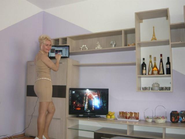 Ghertrude with blonde hair, wearing a tight beige dress, stands on a chair, holding a laptop, looking at a TV with a man's butt on the screen.