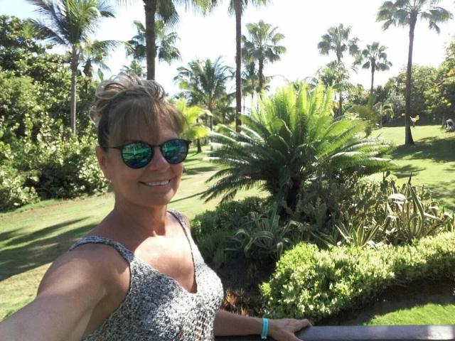 Derlysweet with short blonde hair, wearing reflective green sunglasses, a patterned sleeveless top, and a green bracelet, smiling in a sunny, palm-filled garden. Boobs visible.