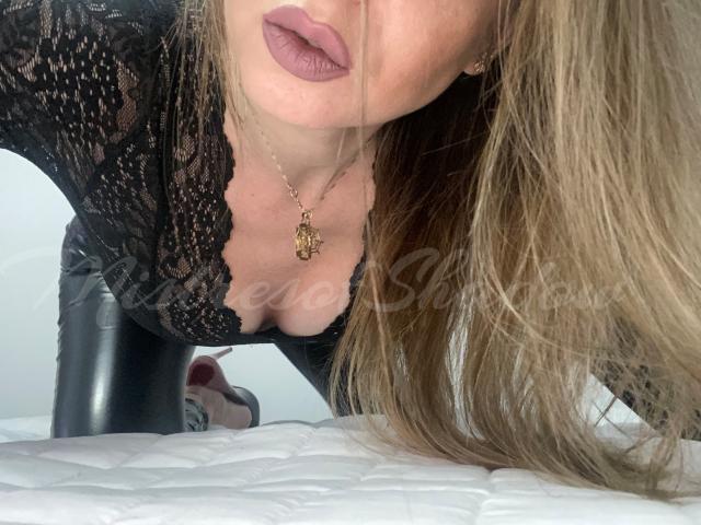 MistressOfShadow with long blonde hair, wearing a black lace top, showing her cleavage and a gold necklace, leaning forward on a white bed. Her pouty purple lips are visible.