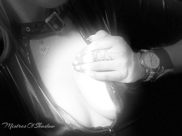 MistressOfShadow's hand with rings touches her bare tits, wearing a leather jacket, collar, and watch. Bright light shines on her chest, highlighting her cleavage.