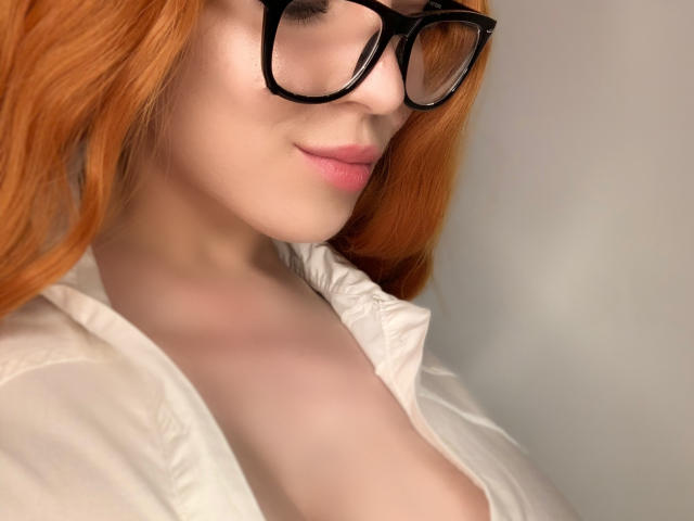 Claire, con i capelli arancioni, gli occhiali neri e le labbra rosa, indossa una camicia bianca, mostrando i suoi seni scoperti, e sorride dolcemente..