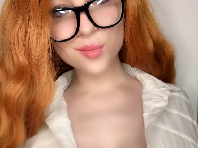 ClaireCream a les cheveux roux, des lunettes noires et des lèvres roses. Elle porte une chemise blanche matelassée qui dévoile sa poitrine, et ses seins sont légèrement visibles. Elle sourit doucement..