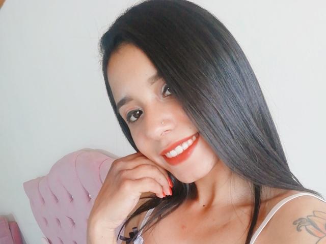 AshleyLover has long black hair, red lipstick, and a nose piercing. She's smiling with red nails, showing a tattoo on her right shoulder. She's wearing a white top.