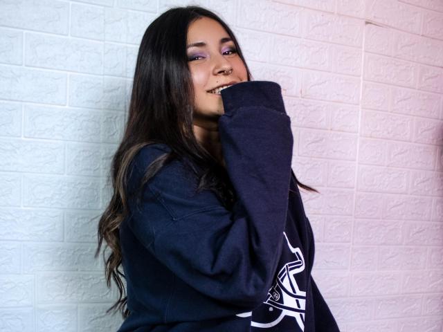MaryJaneHoe with long dark hair, smiling, wearing a navy sweatshirt, purple eyeshadow, and a nose ring, covering her mouth with her hand, looking cute.