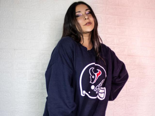 MaryJaneHoe with long dark hair, wearing a loose black shirt with a graphic, standing against a white brick wall, looking slightly to the side with a neutral expression.