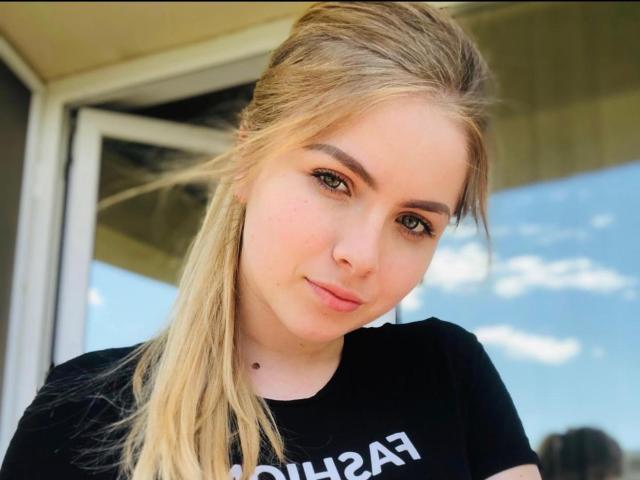 EmaJolie tiene el cabello rubio, ojos verdes y una cara bonita con un pequeño piercing negro en la nariz. Lleva una camisa negra y mira directamente a la cámara con una ligera sonrisa..