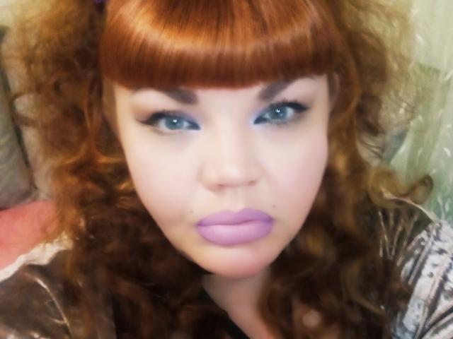 RedBustyX con i capelli rossi ricci, rossetto viola e occhi azzurri, che guarda la fotocamera con un leggero sbuffo. Ha un viso carino e la frangia, e appare sexy..