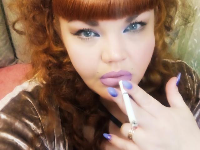 RedBustyX con capelli rossi ricci, occhi azzurri e rossetto viola, che tiene una sigaretta tra le labbra, indossa una camicetta di velluto marrone, con unghie blu curate e un anello..