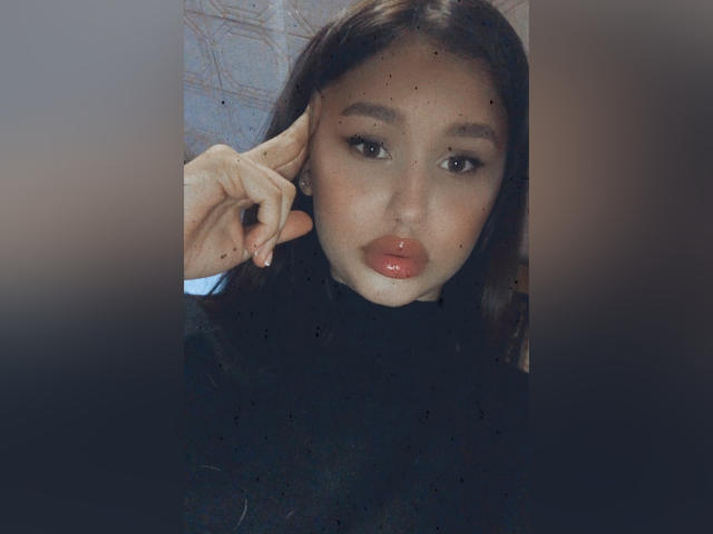 Betty Saunder, con labbra rosse lucide, occhi scuri e lunghi capelli castani, indossa un maglione nero a collo alto e ha una mano che tocca l'orecchio, in un selfie sfocato..