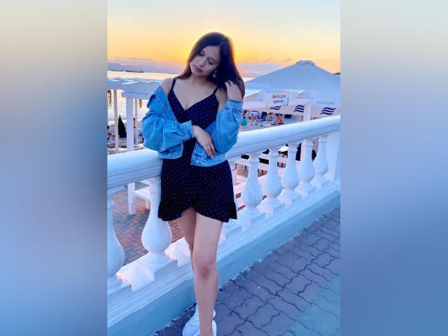 Betty Saunder, con senos de tamaño medio, vestido negro y chaqueta azul, de pie en un balcón al atardecer, mirando hacia abajo, con las manos cerca de la cintura, barandilla blanca, ladrillos azul grisáceos y cielo rosado..