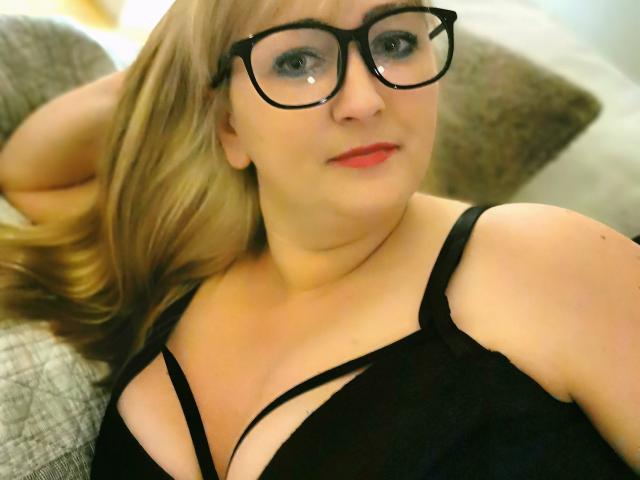 MarinaSweet with glasses, blonde hair, red lipstick, black bra, showing her tits, lying on a couch, looking at the camera with a slight smile.