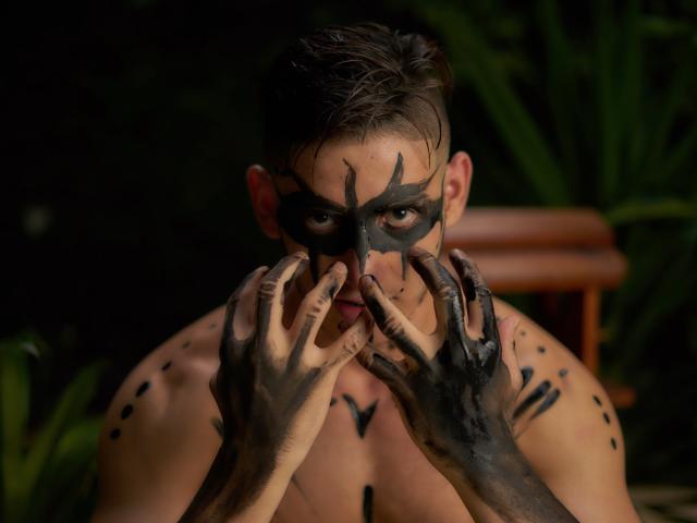 JonathanJeremiah with black face paint, covering his nose with dirty hands, has black spots on his chest and neck, short brown hair, and a serious look, in a dark setting.