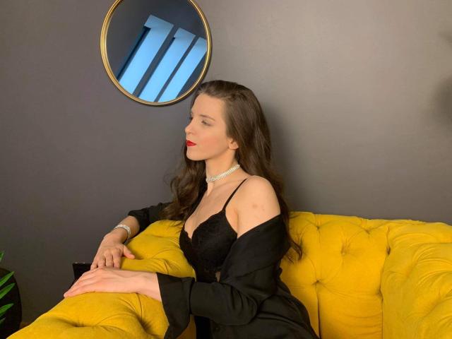 StellaBang with long brown hair, red lipstick, black lace bra, off-shoulder black dress, white choker, sitting on yellow tufted couch, looking to the side, hand on couch.