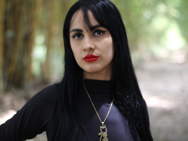 BabeCuteX tiene el cabello negro, lápiz labial rojo y un collar dorado con una cruz. Lleva una blusa negra de manga larga y está mirando directamente a la cámara..