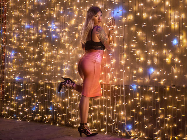 ValerieMartens with long blonde hair, black top, pink leather skirt, and black high heels, standing on one leg, showing her curvy ass and tattooed arm, against a wall of lights.