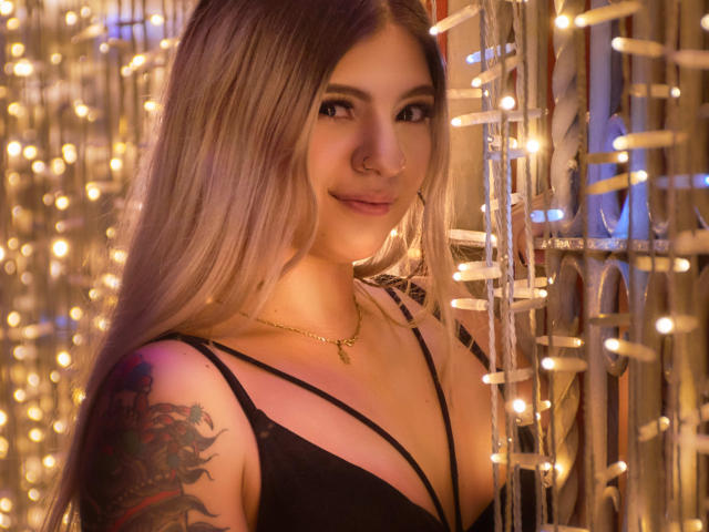 ValerieMartens, avec de longs cheveux blonds, un anneau au nez, un haut noir à bretelles, un tatouage sur l'épaule, un collier, souriante, entourée de guirlandes lumineuses chaudes, debout près d'un mur en fil de fer..