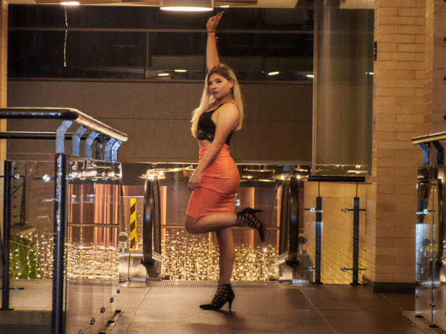 ValerieMartens with long blonde hair, black lace top, orange high-waisted skirt, black strappy heels, and raised arm, standing on one leg, showcasing her curvy ass and tits.