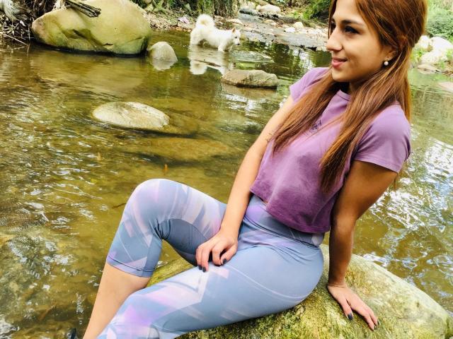 TeequilaCouple69, mit braunen Haaren und einem violetten Oberteil, sitzt auf einem Felsen an einem Bach und trägt enge, violett-blaue Leggings mit Muster, die ihren kurvigen Hintern und ihre kleinen Brüste zeigen..