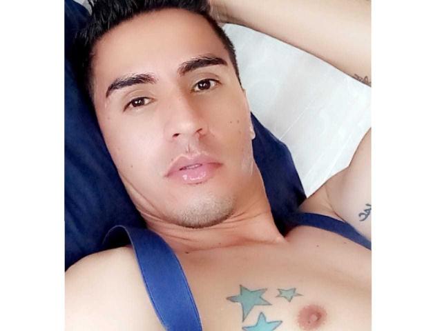 JordanSexyX, met kort zwart haar, bruine ogen en een lichte baard, ligt plat, draagt een blauw tanktopje en heeft een tatoeage van drie groene sterren op zijn borst..