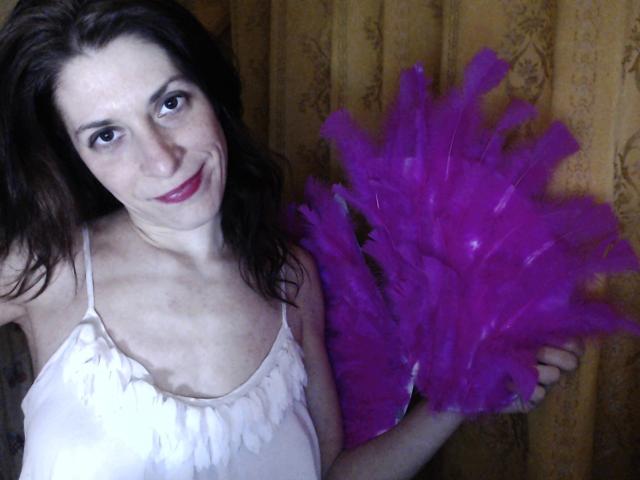 Uma mulher atraente com plumas roxas, um vestido branco e batom vermelho, sorrindo para a câmera. Ela tem cabelos castanhos ondulados e segura as plumas com a mão direita..