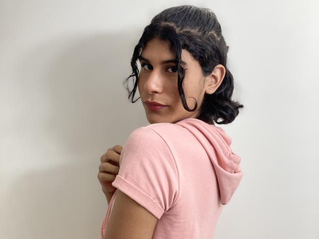 AnnaGrey with wavy black hair, pink hoodie, and side glance, arms crossed, looking over shoulder, medium tits, cute, light skin, subtle smile, white wall background.