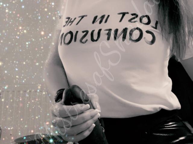 MistressOfShadow with a white t-shirt showing her tits, holding a black dildo with both hands, wearing black shiny pants, and a black wristwatch. Sparkles in the background.