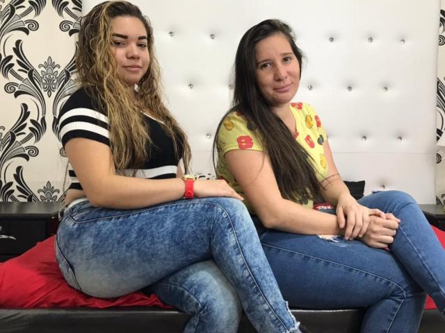 AdrianaYLuna, met krullend blond haar, een zwart-wit gestreepte top en blauwe jeans, zit naast AdrianaYLuna, met lang bruin haar, een gele top met bloemenprint en blauwe jeans, op een bed..