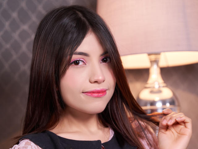 VanityValos met donkerbruin haar, roze eyeliner, roze lippenstift, een zwarte top en een schattige glimlach, die haar haar vasthoudt in de buurt van een zilveren lamp met een beige lampenkap..