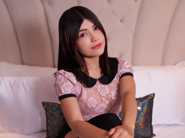 VanityValos avec de longs cheveux noirs, un haut en dentelle rose, un col noir, du fard à paupières rose et du rouge à lèvres rose, assise sur un lit, souriante, les mains sur les genoux, avec un oreiller brillant derrière elle..