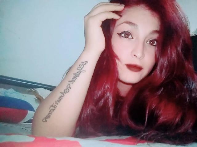 AnastaciaHotBeba con cabello rojo, acostada en la cama, con una mano en la cabeza, un tatuaje en el antebrazo, delineador de ojos, un piercing en la nariz y lápiz labial rojo, mirando a la cámara, con los labios ligeramente entreabiertos..