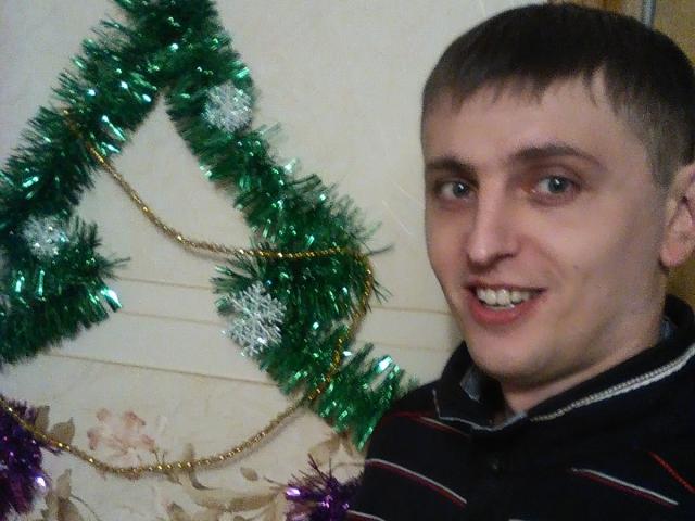 ApollonHot, un tipo un po' eccentrico con i capelli castani corti e un sorriso buffo, indossa una camicia a righe e si trova accanto a un albero di Natale verde decorato con ornamenti viola e argentati..