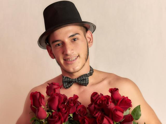 JonathanJeremiah with a black bowtie and hat, holding a bunch of red roses, smiling with a slightly crooked tooth. Shirtless, with a light skin tone, and short hair.