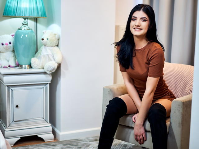 AngelaFay with black hair, brown dress, and black thigh-high boots, sitting on a beige chair, smiling, with white and pink stuffed animals on a white nightstand.