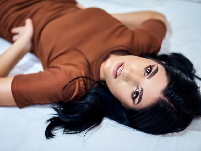 AngelaFay lies on a bed, wearing a brown dress, with black hair and brown eyes, looking up with a slight smile. Her hands are by her side.