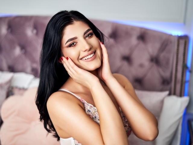 AngelaFay has dark hair, smiling with hands on cheeks, wearing a lacy top, red nails, and looking cute with her tits slightly visible in the soft, pink bedroom.