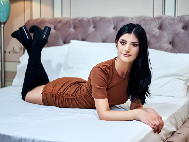 AngelaFay, lying on a bed, wearing a brown dress, black thigh-high boots, and high heels, with long black hair, and a cute, sexy face, looking at the camera.