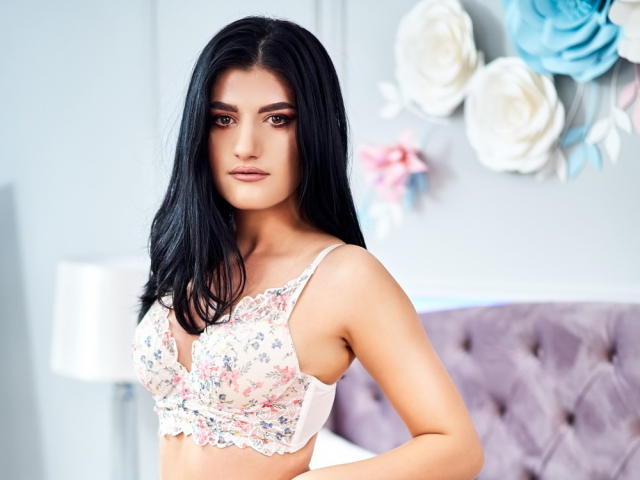 AngelaFay with black hair, wearing a floral bra, showcasing her boobs, standing in a bedroom, looking at the camera with a neutral expression.